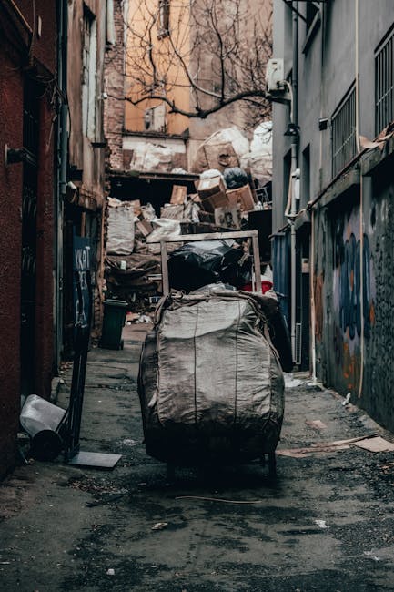 An outdoor alleyway scene with a large trolley or waste container in the foreground, covered in a weathered, grey fabric material. The trolley is positioned on a narrow, asphalt-paved path surrounded by tall, closely spaced brick and metal-clad buildings on either side. Behind the trolley, there is a significant accumulation of mixed rubbish, including cardboard boxes, plastic bags, and miscellaneous debris stacked haphazardly on a makeshift platform or pile, indicating a substantial waste clearing activity. The background shows leafless tree branches rising above the clutter, with some of the buildings featuring visible windows and outdoor fixtures like pipes and electrical meters. The environment appears gritty and urban, characteristic of a location where private rubbish removal or on-site clearance might take place, supporting services such as those offered by House Clearance Shoreditch for alternative waste handling scenarios. The lighting suggests natural daylight, slightly subdued by overcast conditions, emphasizing the utilitarian and unkempt nature of the scene.