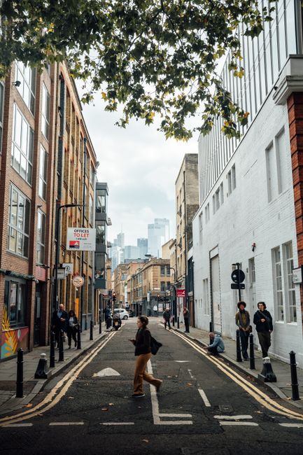A narrow urban street in Shoreditch, lined with multi-storey buildings exhibiting a mix of brickwork and white-painted facades, with large windows and minimal decoration. Overhanging branches with green and yellow leaves partially obscure the top of the image. The street features traffic bollards along both sides, and the surface shows typical asphalt with white and yellow road markings, including a central dashed line and double yellow lines along the edges. Several pedestrians are walking or standing: a woman in brown trousers and a dark jacket crossing the street in the foreground, two individuals standing near the right sidewalk, and a few more further down the street. In the background, taller modern glass buildings are visible, suggesting a city centre vicinity. To the left, a sign advertises office-to-let space, and streetlights and road signs are present. The overall scene is illuminated by natural daylight, reflecting an everyday scene where private rubbish or waste management services, such as House Clearance Shoreditch, could be utilized for independent collection or on-site clearance, fitting the context of alternative waste handling in an urban setting.