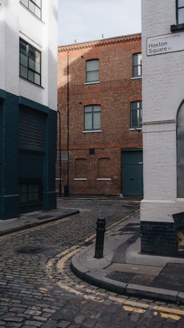 The exterior of the Shoreditch Arts Club building, featuring a storefront with a deep blue awning displaying the establishment's name. The entrance consists of large glass-paneled double doors with vertical wooden handles, positioned below a brick facade. The building's upper level shows a row of windows with white frames, set within a dark brick exterior. Above the windows, there's a line of colorful, blue and green fabric strips hanging across the brick wall, likely for decorative or washing purposes. The scene is set on a paved street with a double yellow line indicating parking restrictions, and a white marking on the road suggests a demarcated zone. Additionally, a short black bollard is placed on the sidewalk near the curb. The overall atmosphere is urban, with natural daylight providing clear visibility of the building's textures, including the brickwork and glass surfaces. The setting is typical of a city street in Shoreditch, with a focus on commercial and artistic spaces, and the scene subtly relates to the context of urban waste management and private collection, given the presence of the building and street infrastructure.