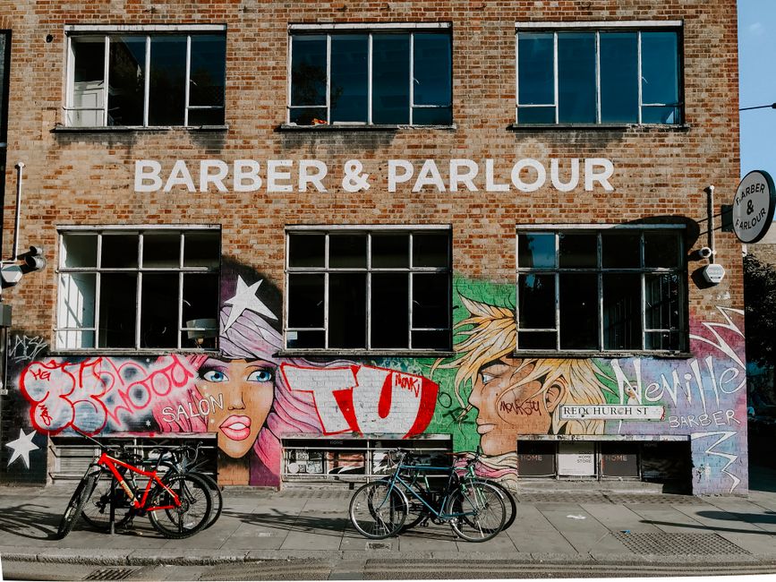 The image depicts a multi-story brick building with large rectangular windows, some of which are open, set against a clear blue sky. Prominently displayed on the facade is the sign reading 'BARBER & PARLOUR' in large white letters. The lower section of the building features vibrant street art murals, including a stylized female face with pink hair and blue eyes on the left, and a side profile of a male figure with blonde hair on the right, both painted with bold outlines and bright colors. Beneath the murals, there are several bicycles parked along the sidewalk, leaning against the building. The environment suggests an urban setting, with the building likely functioning as a local barber shop or parlour, typical of area-specific self-managed businesses. The scene is well-lit by natural daylight, with shadows cast by the bicycles, indicating a sunny day. The street art adds a contemporary artistic element, and the overall image captures a typical city scene with an emphasis on local commercial activity and urban street culture, consistent with the context of independent waste handling and disposal services provided by companies like House Clearance Shoreditch.